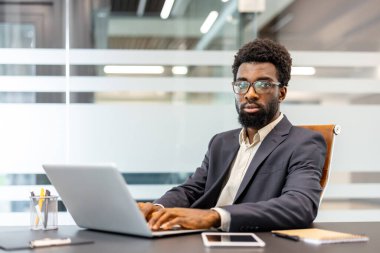 Profesyonel bir ofis masasında dijital çalışmaya odaklanmış, teknolojiyi iş için kullanan ve doğrudan izleyiciye bakan güvenilir bir şirket beyefendisi.