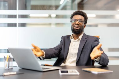 Takım elbiseli endişeli işadamı, modern ofis ortamında bir işyeri teknolojisi sorunuyla boğuşurken stres ve hayal kırıklığı göstererek dizüstü bilgisayarında hayal kırıklığı içinde el kol hareketi yapıyor.