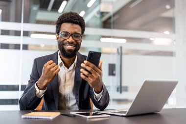 Genç işadamı modern ofiste yumruk tokuşturuyor, akıllı telefonunu kontrol ederken aynı zamanda bilgisayar ve tabletin ortasında başarılı dijital haberler ve başarılara gülümsüyor.