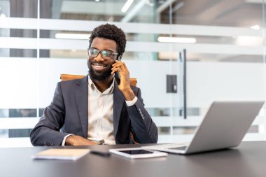 Kendine güvenen bir iş adamı, modern bir ofiste masasında çalışırken telefon konuşmasından zevk alıyor. İletişim, ağ ve başarılı bir kariyer yönetimi sergiliyor.
