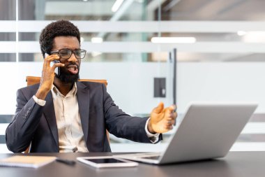 Takım elbiseli ve gözlüklü profesyonel bir adam, ofis masasındaki akıllı telefonuyla konuşur, bilgisayarına ve tabletine el kol hareketi yapar, bir yandan da iş görüşmesine ve uzaktan görüşmeye konsantre olur.