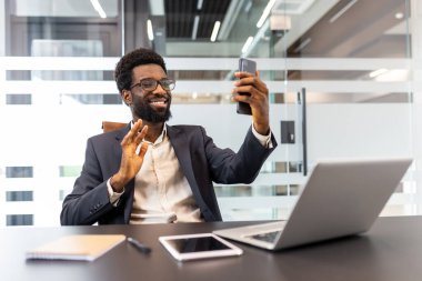 Profesyonel çalışma alanından sanal toplantıya ya da video konferansına katılan gülümseyen Afrikalı iş adamı, cep telefonu veya müşterileriyle uzaktan iletişim kuruyor.