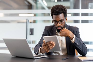 Afrika kökenli Amerikalı iş adamı modern ofis masasında oturuyor, gözlük takıyor ve resmi bir takım elbise giyiyor, profesyonel bir işe odaklanırken dijital tablet kullanıyor.