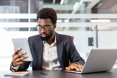 Modern bir ofiste çalışan profesyonel işadamı, bir dizüstü bilgisayar ve dijital tabletle birden fazla iş yapıyor, verimlilik gösteriyor ve kurumsal görevlerine bağlılığını gösteriyor.