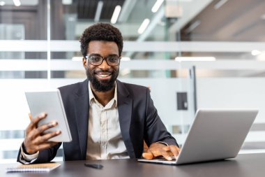 Dijital cihazlarla verimli bir şekilde çalışan, görevleri yönetirken kameraya bakan ve gülümseyen ve çağdaş bir çalışma alanına bağlanan, başarı ve yeniliği somutlaştıran kendine güvenen iş adamı