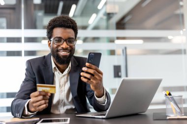 Gülümseyen adam, ofis masasında elinde altın bir kredi kartı ve akıllı telefon tutarak, e-ticaret, bankacılık ve modern dijital işlemleri gösteren güvenli bir cep telefonu ödemesini tamamlıyor.