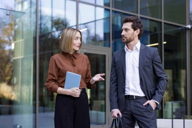 Bir tablet tutan ve takım elbiseli bir erkek meslektaşıyla iletişim kuran iş kadını, ikisi de modern bir şirket binasının önünde çalışma stratejileri hakkında açık havada konuşuyordu.
