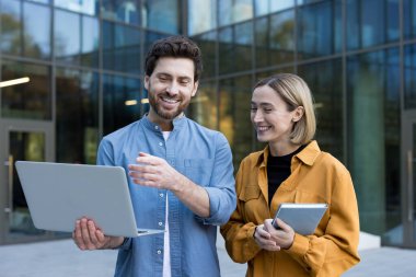 Dışarıda duran iş arkadaşları, elinde dizüstü bilgisayar olan bir adam ve tabletli bir kadın gülümserken el kol hareketi yaparak ortak fikirleri ve takım çalışmasını tartışıyor.