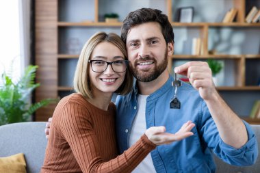 Kanepede oturan ve elinde yeni ev anahtarlarıyla gülümseyen genç bir çift. İlk evlerine ya da dairelerine taşınmalarını kutluyorlar. Gayrimenkul yatırımını ve aile geleceğini temsil ediyorlar.