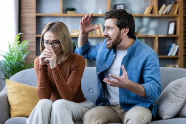 İlişki sorunları yaşayan bir çift, sinirli ve öfkeli bir adam üzgün ve üzgün karısına bağırıyor, koltukta oturuyor, duygusal sıkıntı, taciz ve üzüntü yaratıyor.