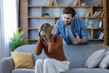 Bir kadın kulaklarını kapatırken, bir erkek evdeki çatışmaları, stresi ve ilişki sorunlarını resmeden hararetli bir aile içi tartışma sırasında kızgın bir şekilde arkasından el kol hareketi yapıyor.