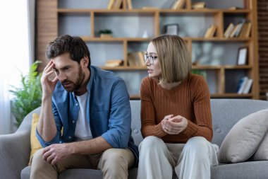 Oturma odasındaki kanepede tartıştıktan sonra gergin bir sessizlik içinde bir çift, kadın konuşurken başını tutan bir erkek, stres, iletişim bozukluğu ve ilişki gerginliği tasvir ediyor.