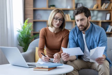 Genç çift, evdeki bir koltukta oturup, belgeleri tutarken ve finansal planlama için dizüstü bilgisayar kullanırken, ev ekonomisi, hesaplama ve bütçe hesaplamaları yapıyor.