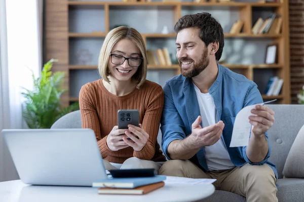 Mutlu genç çift evde bir kanepede otururken gülümsüyor, akıllı telefon ve dizüstü bilgisayarla faturaları ödüyor, finans işlerini yürütüyor ve ev harcamalarını gözden geçiriyorlar.