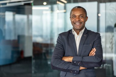 Arkadaş canlısı bir gülümseme ve kollar kavuşturulmuş, çağdaş bir kurumsal ortamda duran, başarıyı ve liderliği yansıtan kendinden emin bir iş adamı.