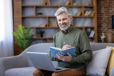 Olgun bir adam gülümsüyor, koltukta rahatça oturuyor, dizüstü bilgisayardan çalışırken bir defter ve yazı tutuyor, esnek uzaktan kumandanın özünü yakalıyor.