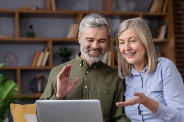 Bir dizüstü bilgisayardaki video görüşmesi sırasında gülümseyen ve el sallayan mutlu yaşlı çift, aile veya arkadaşlarla iletişim kuruyor ve rahat evlerinden modern iletişim teknolojisini kucaklıyorlar.