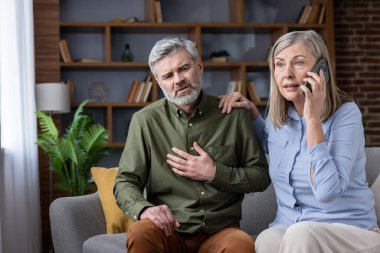 Yaşlı adam ani kalp krizi ya da göğüs ağrısından muzdarip, göğsünü tutuyor, bu sırada endişeli karısı akıllı telefondan acil servisleri arayıp tıbbi yardım istiyor.