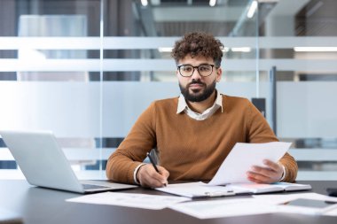 Gözlüklü ve sakallı genç bir adam modern bir ofiste masa başında oturuyor, evrak işlerine konsantre oluyor ve dizüstü bilgisayar kullanıyor, iş analizini ve profesyonel işi temsil ediyor.