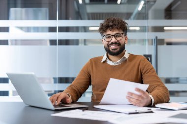Bilgisayarda çalışırken kameraya gülümseyen ve elinde evrak tutarak çağdaş bir ofis ortamında masa başında oturan genç profesyonel bir adam.