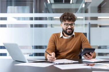 Finans üzerinde çalışırken gülümseyen bir adam, elinde bir hesap makinesi tutarken ve kalemle kağıda yazarken, belgelerle çevrili ve modern ofis ortamında bir dizüstü bilgisayarla