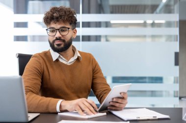 Genç işadamı, üretkenlik ve çoklu görevi vurgulayarak, modern ofis masasında dizüstü bilgisayar kullanırken dijital tablet ve bir not defterine yazı yazarak verimli bir iş yaptı.