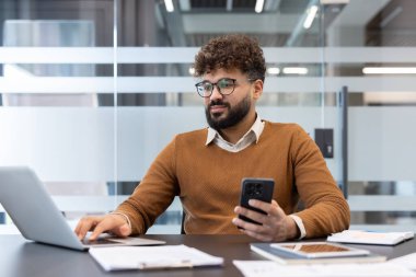Hintli işadamı cep telefonu tutarken bilgisayarında çalışıyor, çağdaş bir şirket ortamında uzaktan çalışma, çoklu görev ve dijital iletişimi temsil ediyor.