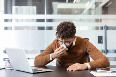 Kıvırcık saçlı, gözlüklü, burnunu mendile silen bir adam modern bir ofiste, çalışma masasında çalışırken, işyeri sağlığı ve hastalığını resmediyor.