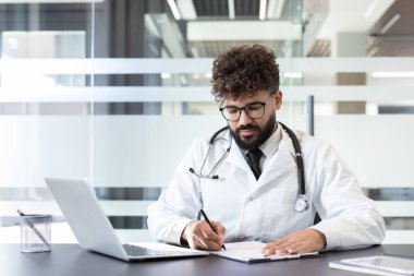 Kıvırcık saçlı, sakallı, gözlüklü ve steteskoplu beyaz önlüklü genç bir erkek doktor, bir masada oturuyor ve modern bir klinikte ya da hastane ofisinde bir panoya yazı yazıyor.