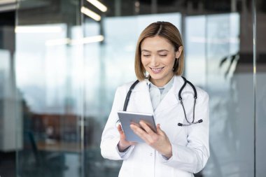 Beyaz önlüklü ve steteskoplu kadın doktor, dijital tabletle etkileşim halindeyken gülümsüyor, klinikte dijital sağlık, teletıp ve modern tıbbi teknolojiyi temsil ediyor.