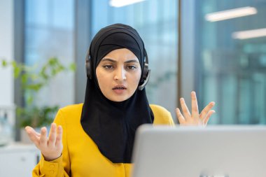 Başörtüsü takan Müslüman bir kadın, modern bir ofiste bilgisayar görüntülerinde kafası karışmış ve hayal kırıklığına uğramış bir halde uzaktan iletişimle mücadele ediyor.