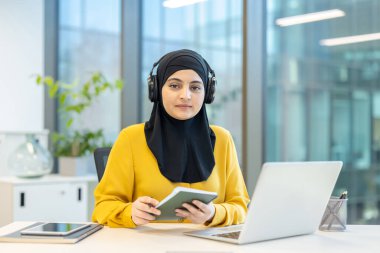 Siyah tesettürlü ve sarı tişörtlü Müslüman bir kadın, bir ofis masasında kulaklıklarla oturuyor, elinde bir dizüstü bilgisayarla kameraya bakıyor.