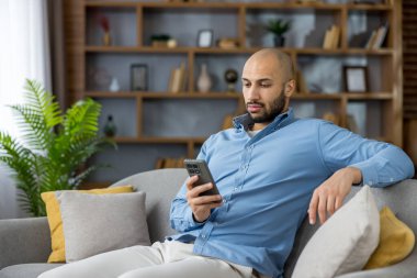 Modern bir oturma odasında gri bir koltukta dinlenen sakallı bir adam, elinde akıllı bir telefon ve kaydırmalı sosyal medya, evde otururken içeriği ve mesajları karıştırıyor ve rahat ediyor.