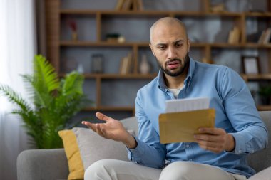 Evdeki kanepede oturan bir adam, bir mektup açıp kafası karışmış veya endişeli görünüyor, kötü haberler okuyor, beklenmedik bir fatura ya da strese yol açan bir mali tebligat.
