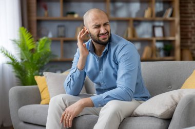Kulak ağrısından muzdarip bir adam, yüzünü ekşiterek kulaklarını ovuyor, evde bir kanepede oturmuş, sağlık sorunları ve işitme sorunu ile boğuşuyor.