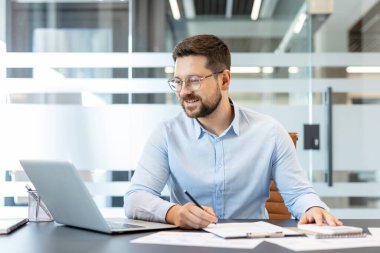 Gözlüklü ve mavi gömlekli genç bir adam modern bir ofis masasında oturuyor, bir dizüstü bilgisayar kullanıyor ve kağıt üzerinde yazıyor, kendini işine adamış ve yoğun çalışıyor.