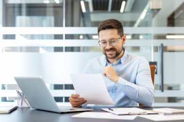 Finans belgelerini inceleyen, veri hesaplayan ya da şirket iş yerinde dizüstü bilgisayarla çalışırken iş raporları okuyan gözlüklü profesyonel bir adam.
