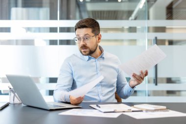Stresli işadamı, modern bir ofis çalışma alanında beklenmedik borçlar, muhasebe hataları ve mali krizle karşı karşıya kalırken, elinde belge ve evrak tutarak dizüstü bilgisayarına şok oldu.