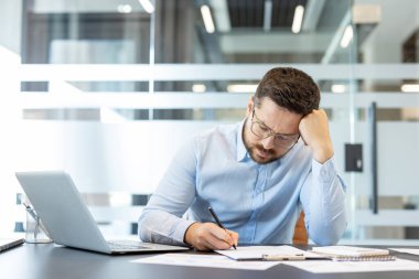 Stresli işadamı hayal kırıklığına uğramış ve bunalmış hissediyor, dizüstü bilgisayar ve belgelerle uğraşırken başını tutuyor, bir şirket ofisinde baş ağrısı ya da tükenme yaşıyor