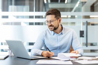 Genç sakallı iş adamı, parlak, modern bir masada gözlüklü, laptopa ve evrak işlerine odaklanırken kendine güvenen, üretken profesyonel planlama raporları yazan ve verileri analiz eden.