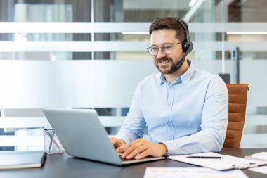 Genç adam, mikrofon ve açık mavi gömlekli bir kulaklık takıyor modern bir ofis masasında oturuyor, dizüstü bilgisayarda yazıyor, müşteri hizmetleri ve destek desteği sağlıyor.