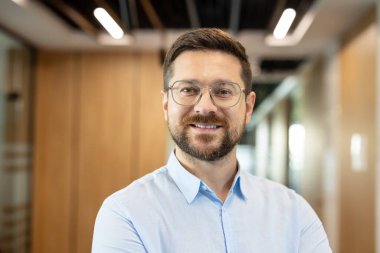 Kendine güvenen profesyonel bir erkek gözlük takıyor ve açık mavi gömlek giyiyor. Kurumsal bir ortamda liderlik ve başarıyı temsil ediyor.