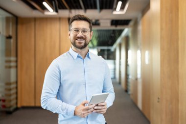 Dijital tablet tutarken gülümseyen kendine güvenen işadamı, bir ofis koridoru boyunca duruyor, işyerinde profesyonelliği, başarıyı ve teknolojiyi temsil ediyor.