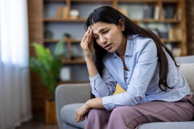 Koyu renk saçlı genç bir kadın evdeki koltukta oturuyor, alnını tutuyor ve migren ya da şiddetli bir baş ağrısı yüzünden irkiliyor, yorgun, stresli ve duygusal olarak tükenmiş.