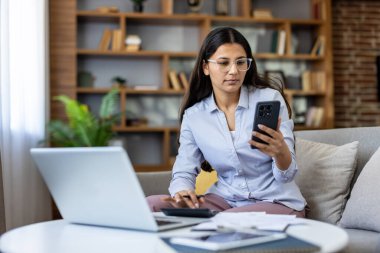 İç işlerindeki koltukta oturan genç bir kadın bilgisayar, akıllı telefon ve hesap makinesiyle birden fazla iş yapıyor. Faturaları ve bütçe hesaplarını gözden geçiriyor. Uzaktan çalışıyor ve ev işleriyle uğraşıyor.