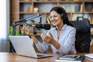 Genç bir kadın gülümsüyor ve profesyonel bir mikrofona kulaklıkla konuşuyor, modern bir ev stüdyosunda canlı yayın yapıyor, kendinden emin bir yaratıcı ve sunucu.