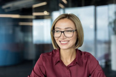 Kısa sarı saçlı ve gözlüklü profesyonel iş kadını kendinden emin bir şekilde gülümsüyor ve başka tarafa bakıyor, modern ofis ortamında duruyor, başarıyı ve liderliği temsil ediyor.