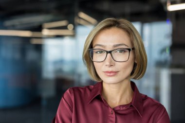 Kendine güveni tam bir iş kadını şık gözlükler takıyor ve bordo bir gömlek giyiyor. Çağdaş ofis ortamında kameraya bakarken profesyonellik ve liderlik tasvir ediyor.