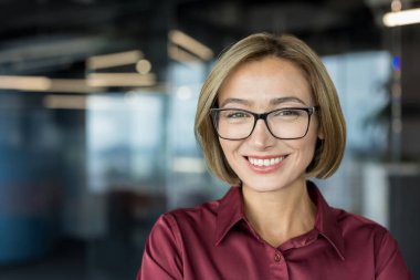 Kendine güveni tam bir iş kadını, gözlük takıyor, bordo bir gömlek giyiyor, modern ofis ortamında profesyonellik ve liderlik taslıyor.