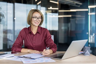 Young businesswoman with glasses smiling confidently while working on paperwork at a desk with a laptop and tablet, focusing on strategy and planning in a professional corporate environment